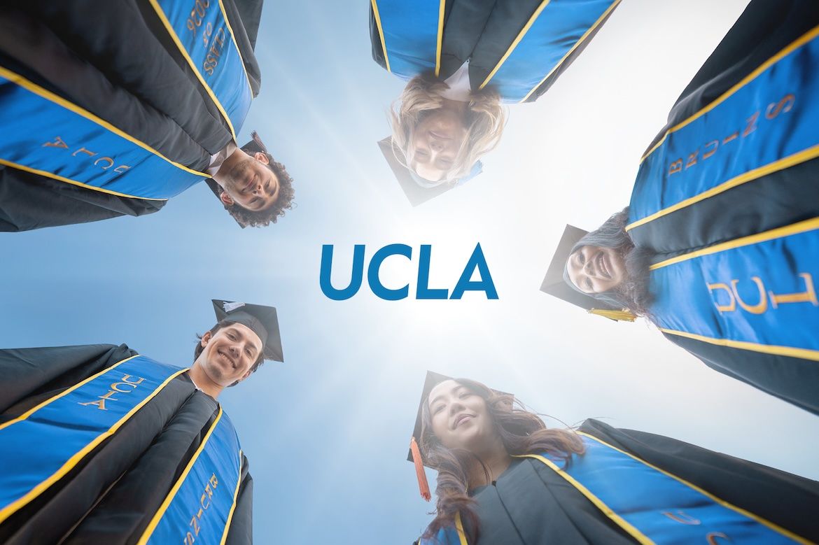 Five graduates in black caps and gowns with blue-and-gold stoles form a circle, viewed from below against a bright sky, surrounding the centered UCLA logo in a celebratory commencement scene.