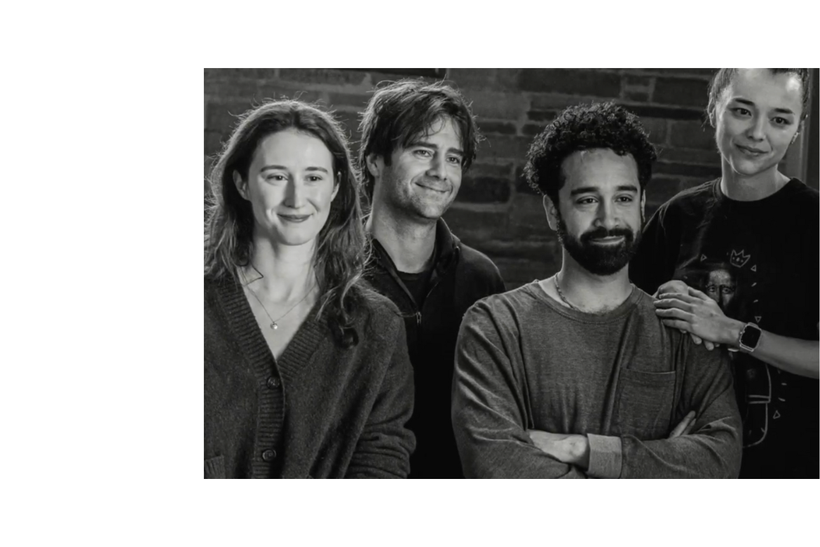 A black-and-white photo shows four adults standing closely together in front of a brick wall, smiling softly at the camera. They are dressed casually, with relaxed postures that suggest a friendly, collaborative group setting.