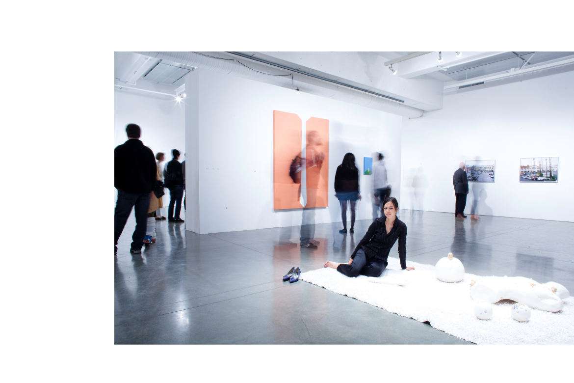 Contemporary art gallery with visitors viewing wall-mounted works. In the foreground, a woman sits on a white rug beside sculptural objects, with shoes placed nearby.