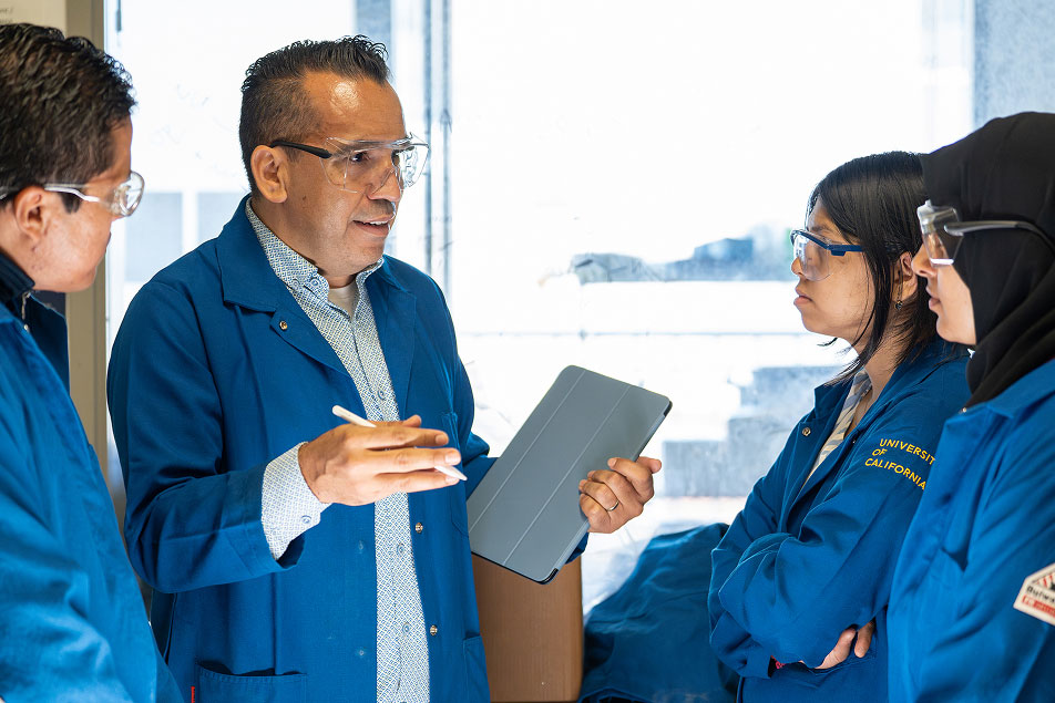 Professor Osvaldo Gutierrez holding pen and pad, talks to three others who are also in blue lab coats.